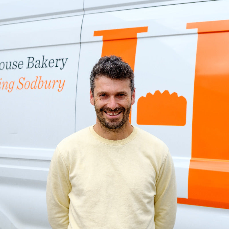 A smiling man with short dark hair and a beard stands in front of a white bakery van with large orange lettering and a bread icon. The text on the van includes 'House Bakery' and 'Sodbury'.