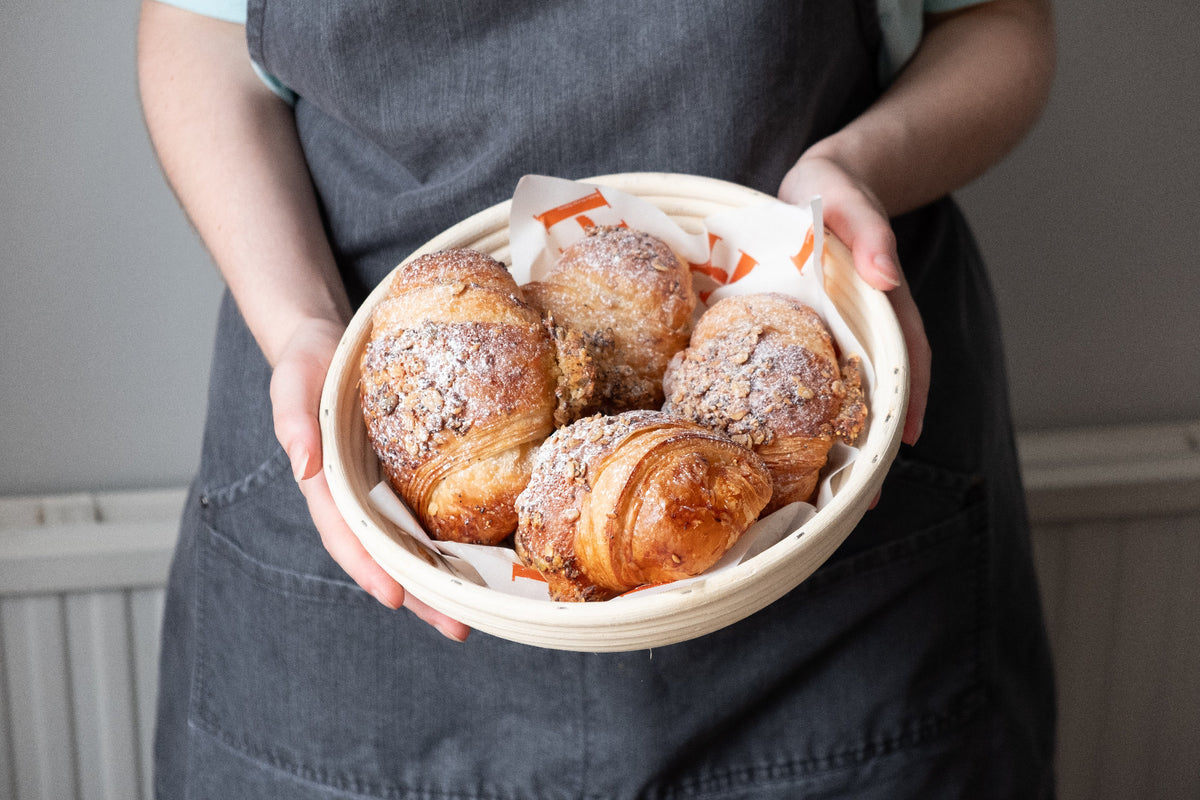 a basket of freshly golden crispt croissants topped with oats and icing sugar, held by a person in an apron.