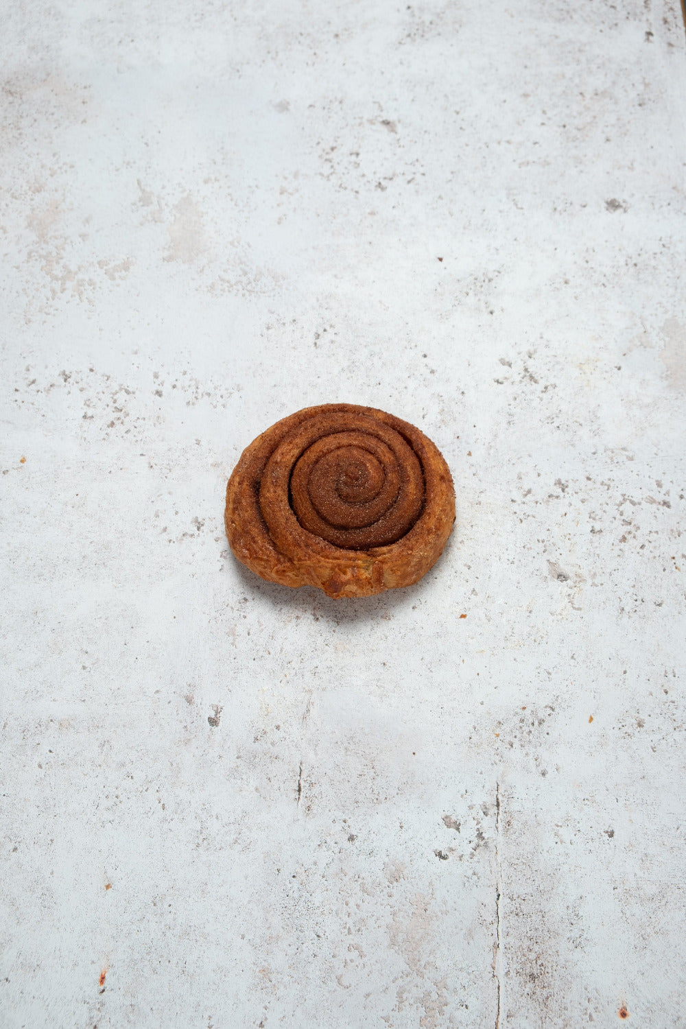 A single cinnamon roll or spiral pastry centered on a light, textured off-white surface.