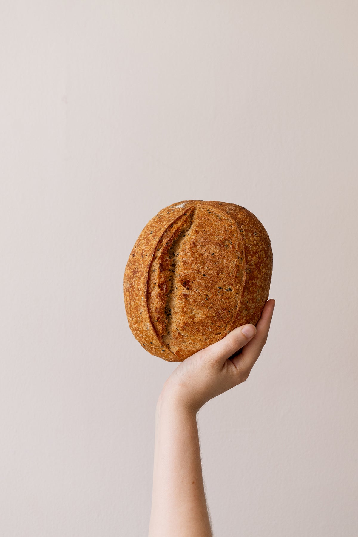 A golden, seeded, round loaf being held by a hand up against a grey background/