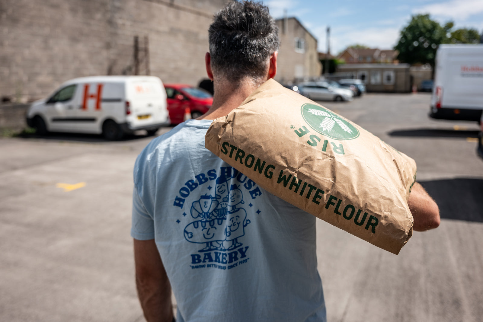 Person wearing a t-shirt with a bakery logo and holding a flour bag in an outdoor setting.