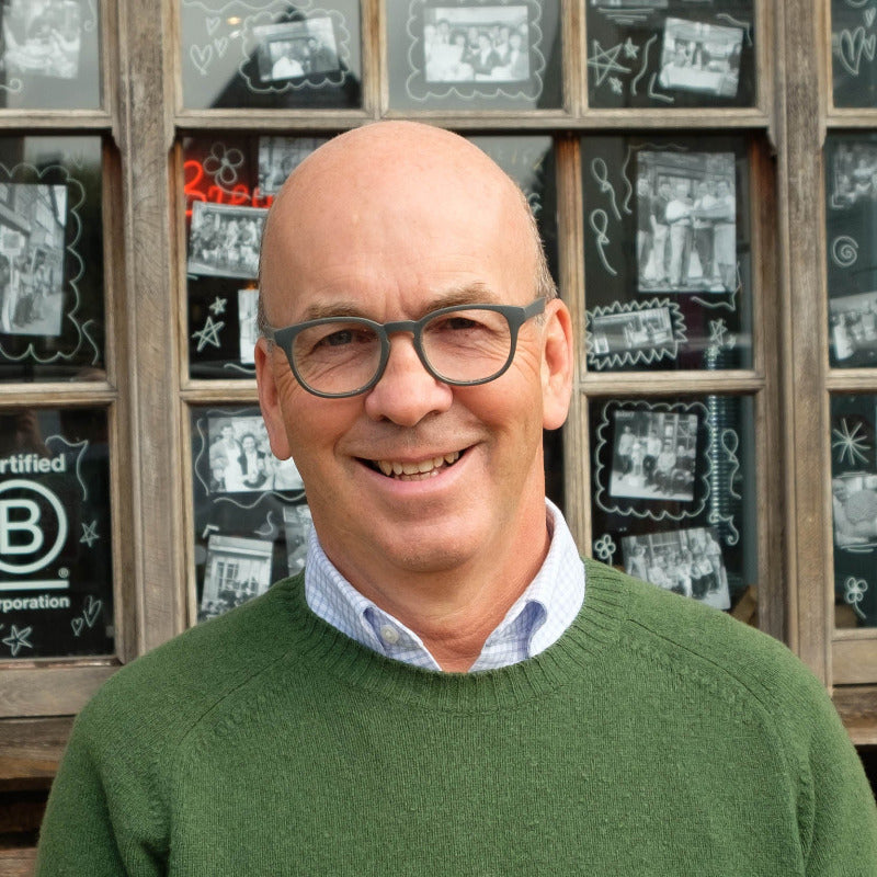 A smiling bald man with glasses, wearing a green crewneck sweater over a light blue collared shirt, standing in front of a rustic wooden window display filled with framed photos.